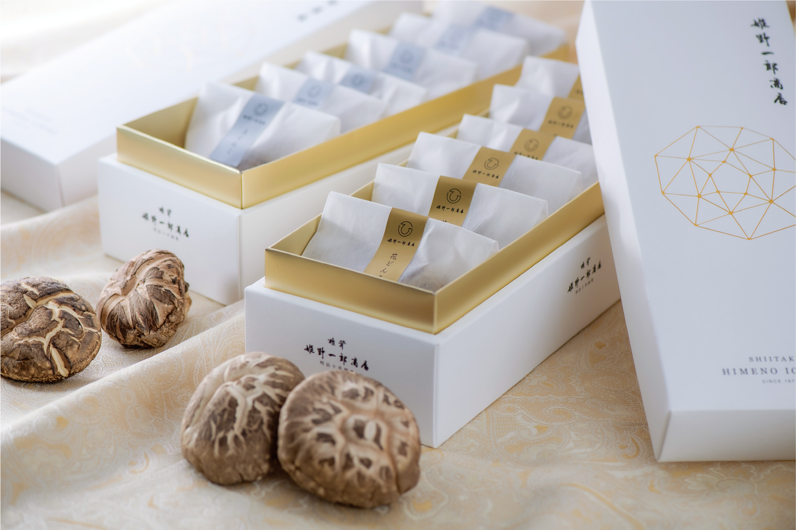 姫野一郎商店の干ししいたけギフトセット。白い上品な箱に「花どんこ」「どんこ」と書かれた帯が付いた個包装のしいたけが並び、箱のそばには乾燥しいたけが置かれている。箱にはリニューアル後のロゴと幾何学模様が金色であしらわれている。
