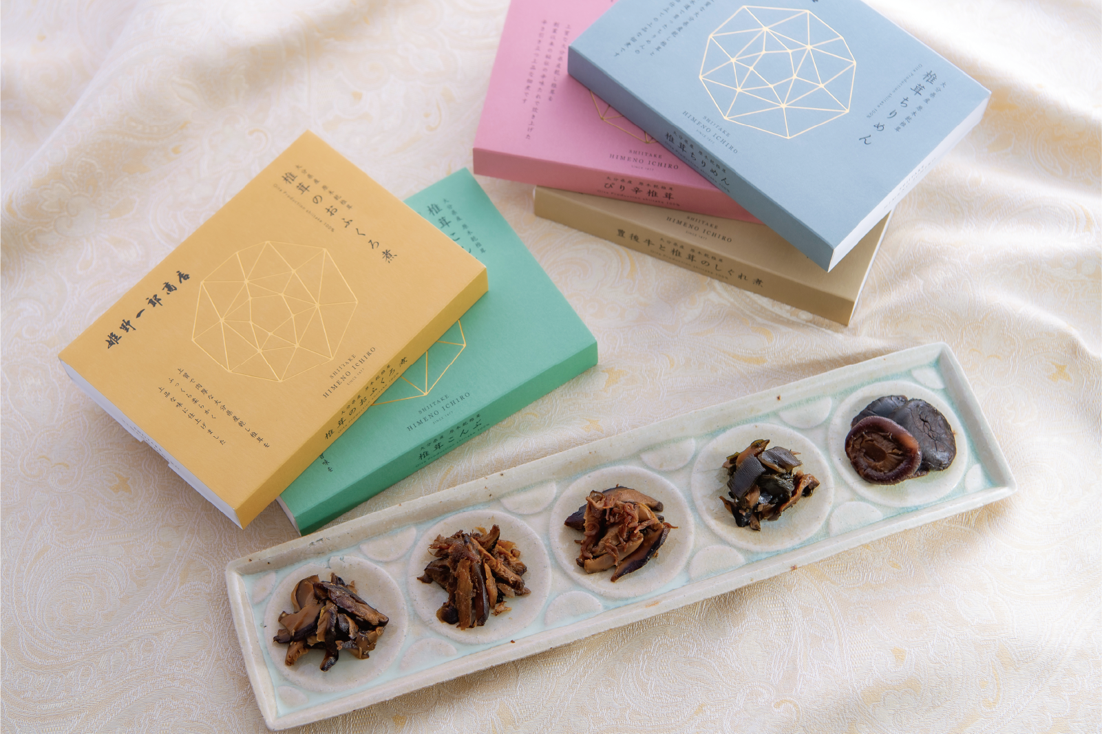姫野一郎商店の椎茸加工品。様々な種類の椎茸の佃煮や煮物が白い長皿に盛り付けられ、その周りにカラフルな箱パッケージ（黄色、緑、水色、ピンク）が置かれている。