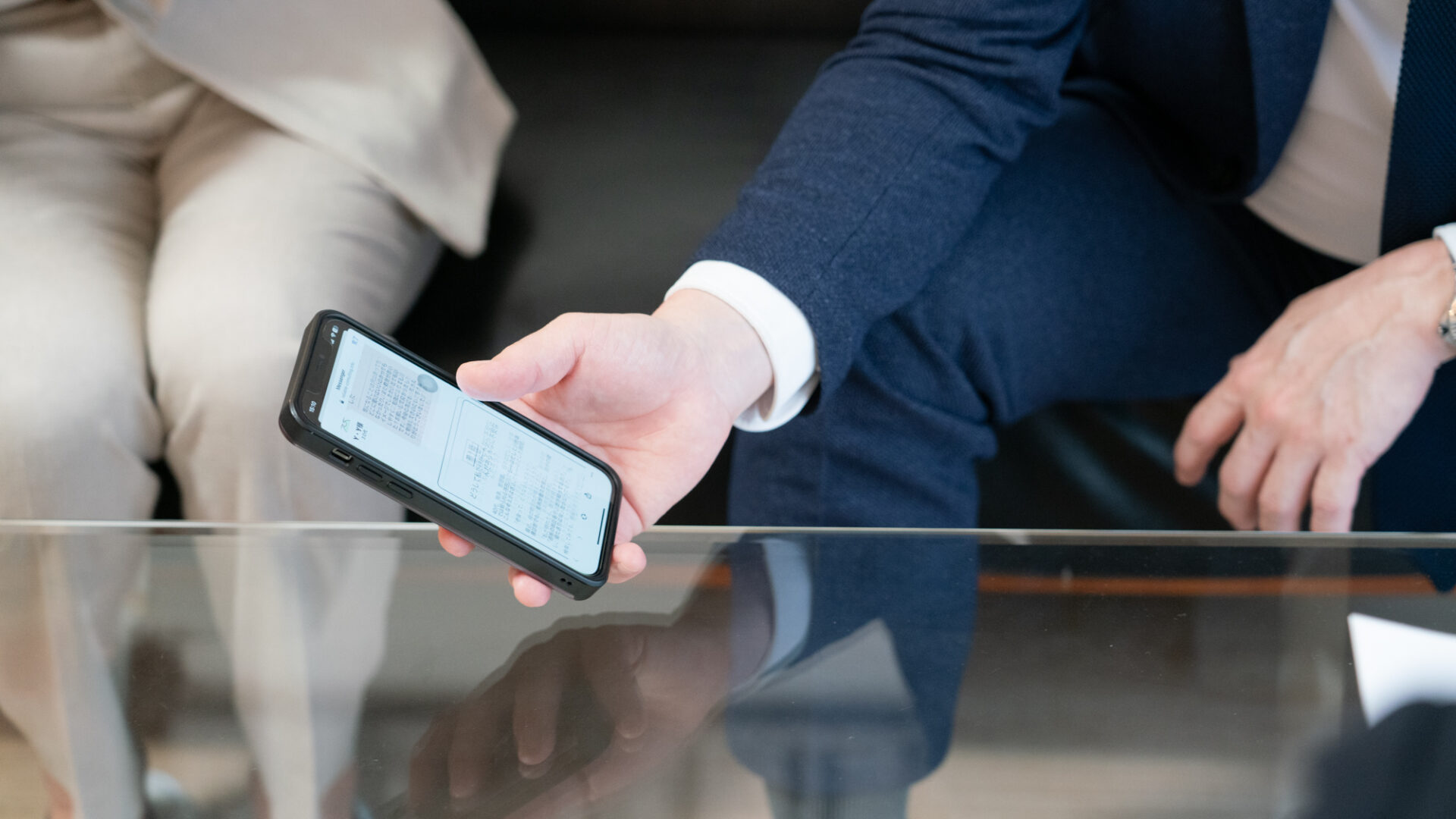 スーツ姿の男性がスマートフォンの画面を相手に見せながら、電子的な資料や情報を提示している場面