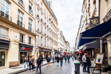 パリの商店街で多くの歩行者が行き交う石畳の通りと両側に並ぶ店舗やカフェ