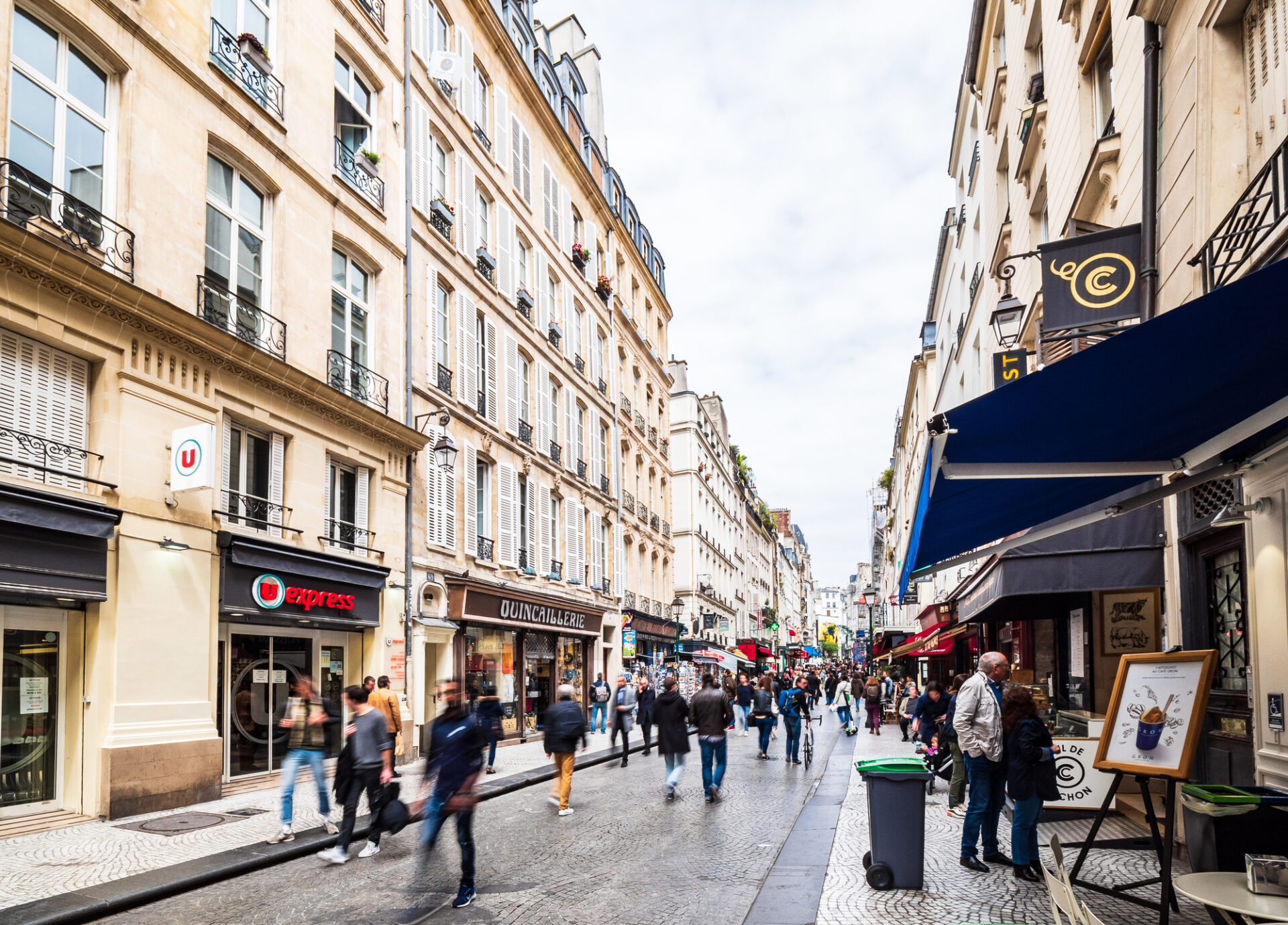 パリの商店街で多くの歩行者が行き交う石畳の通りと両側に並ぶ店舗やカフェ